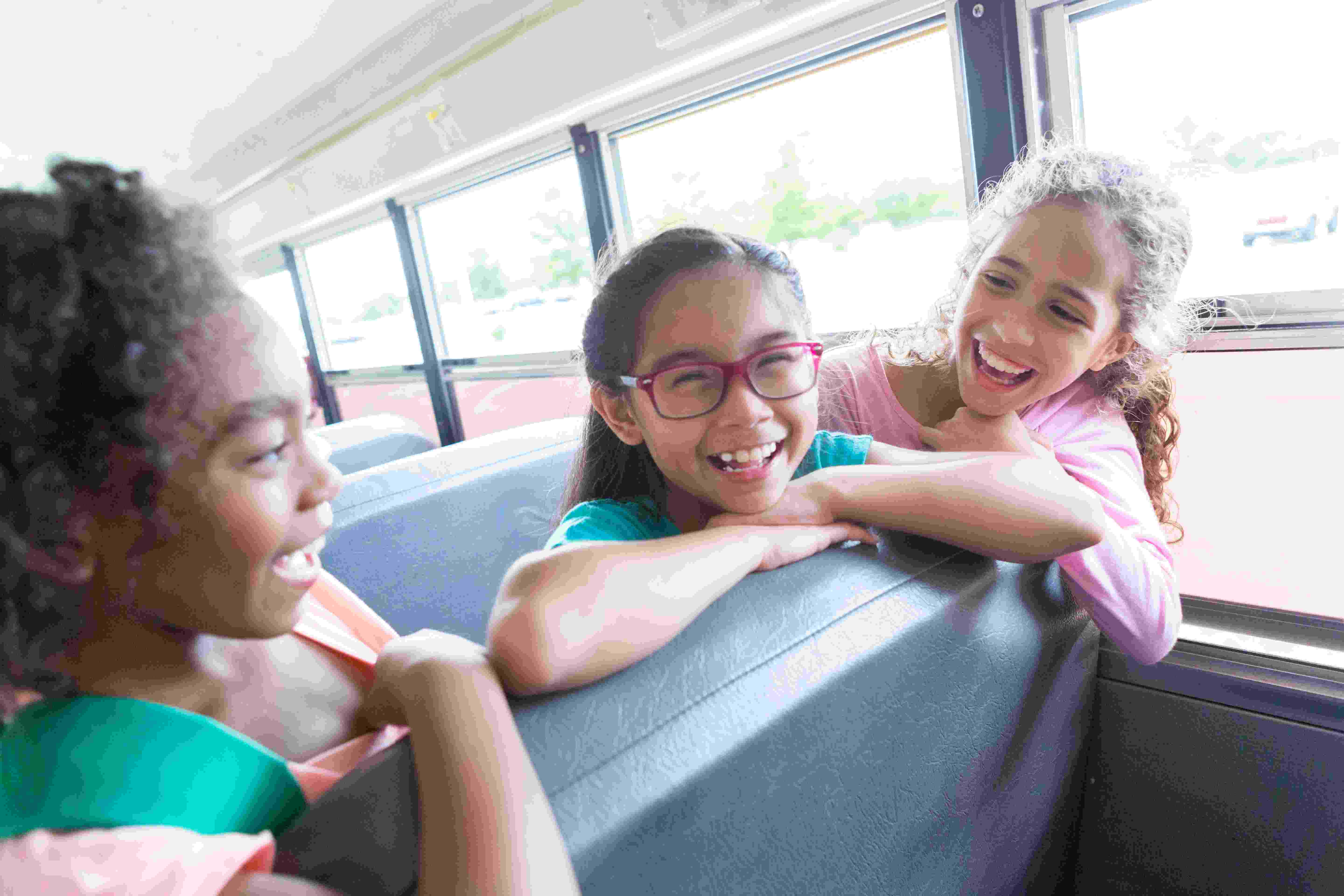 900966080 Diverse elementary school girls laugh with one another as they ride to school on a school bus.